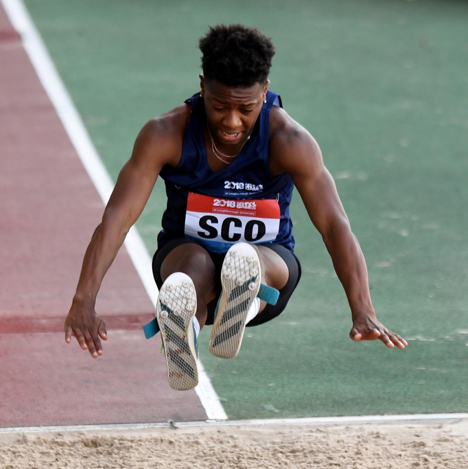 A long jumper wearing a navy blue uniform and a SCO bib is mid-air above a sandpit, arms spread out and legs extended forward, about to land during a track and field event.