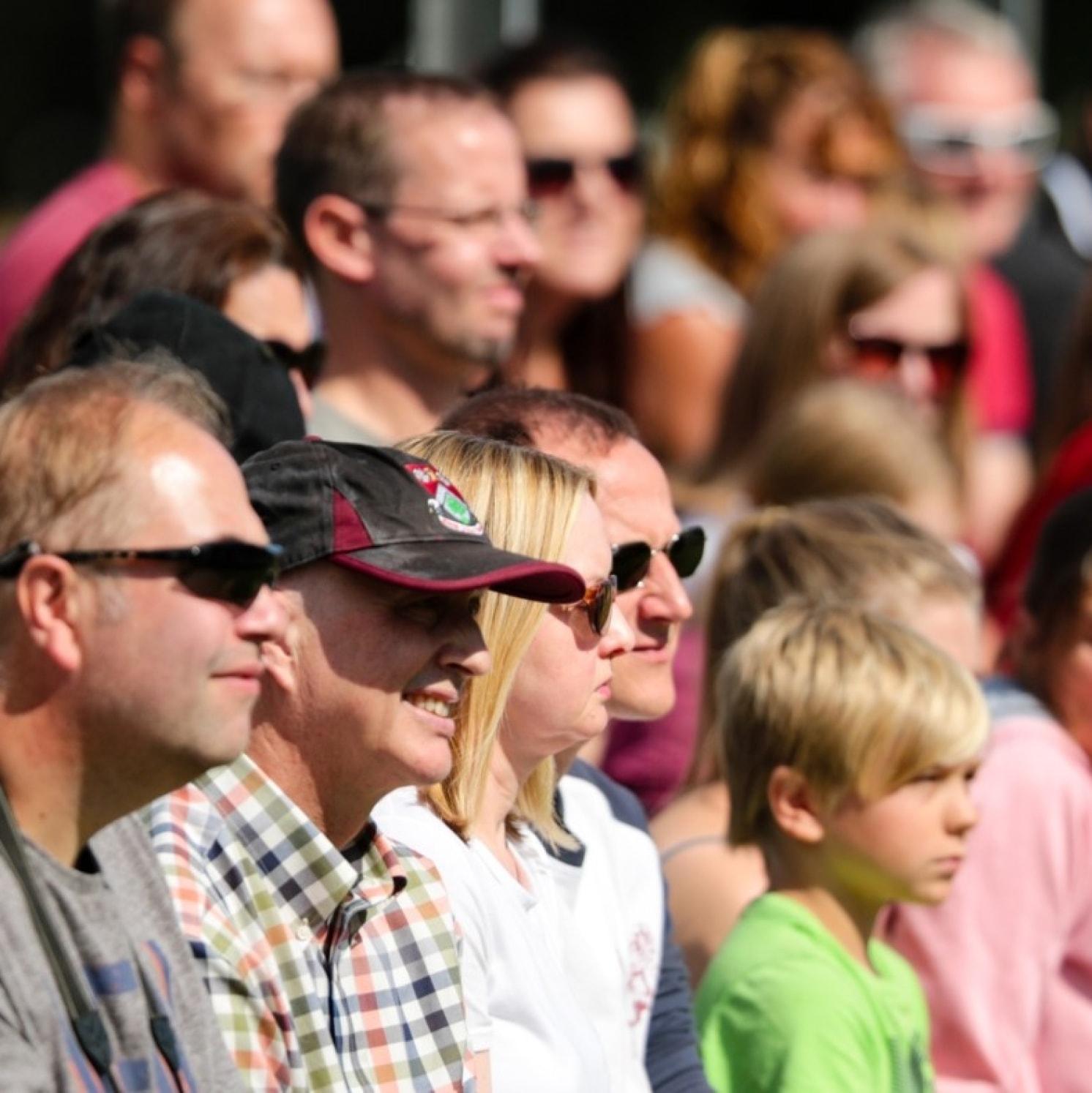 A crowd of people, including adults and a young boy, sit outdoors in bright sunlight, watching an event. Most are wearing sunglasses and casual clothing, and they appear focused on what they are viewing.