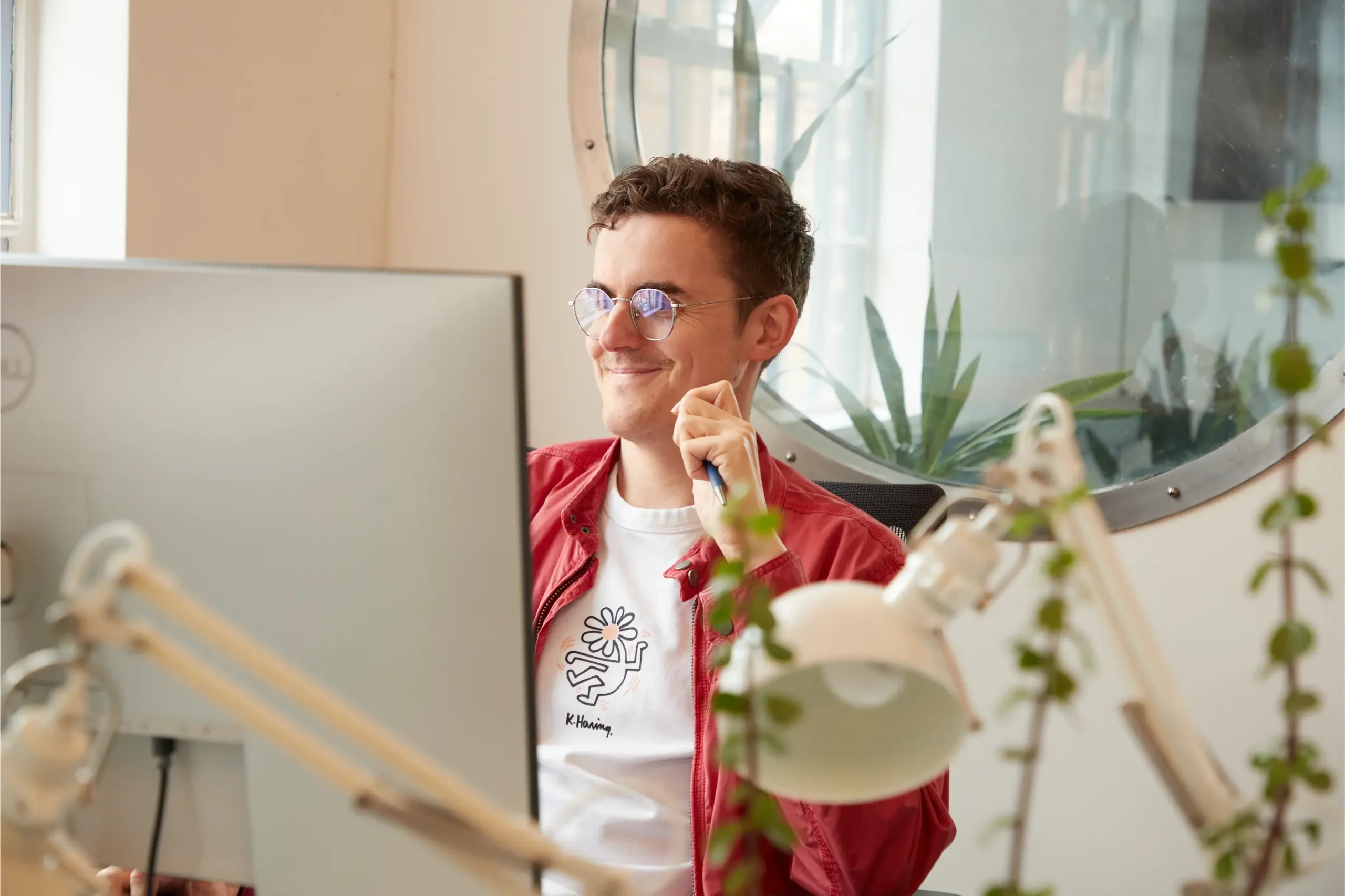A young man wearing glasses and a red shirt smiles whilst sitting at a desk with a computer monitor, holding a pen, surrounded by plants and office lamps in a bright, modern office space.
