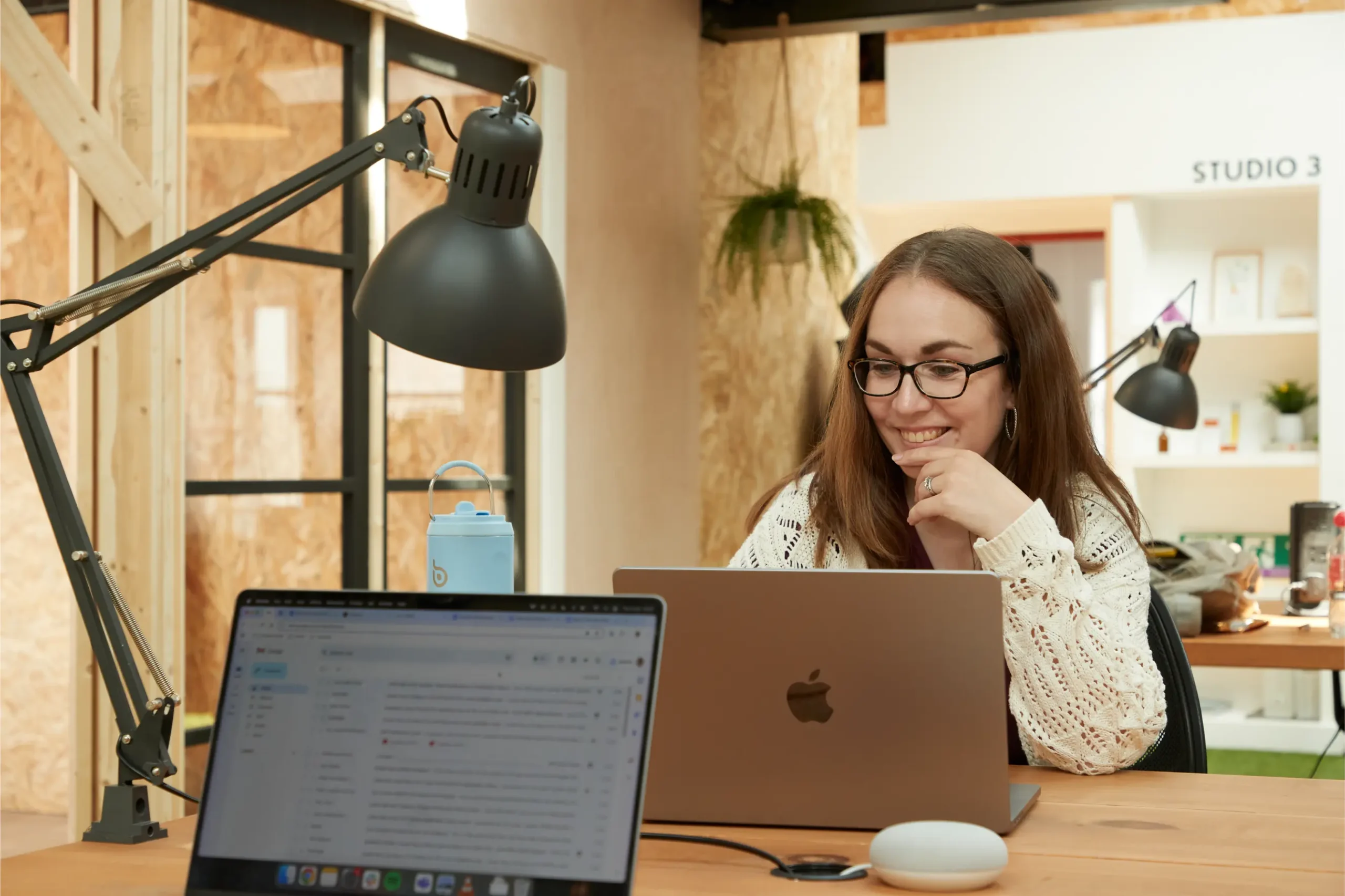 A woman with long brown hair and glasses smiles whilst working on a laptop at a desk in a modern office, with a lamp, water bottle, and another open laptop visible in the foreground.