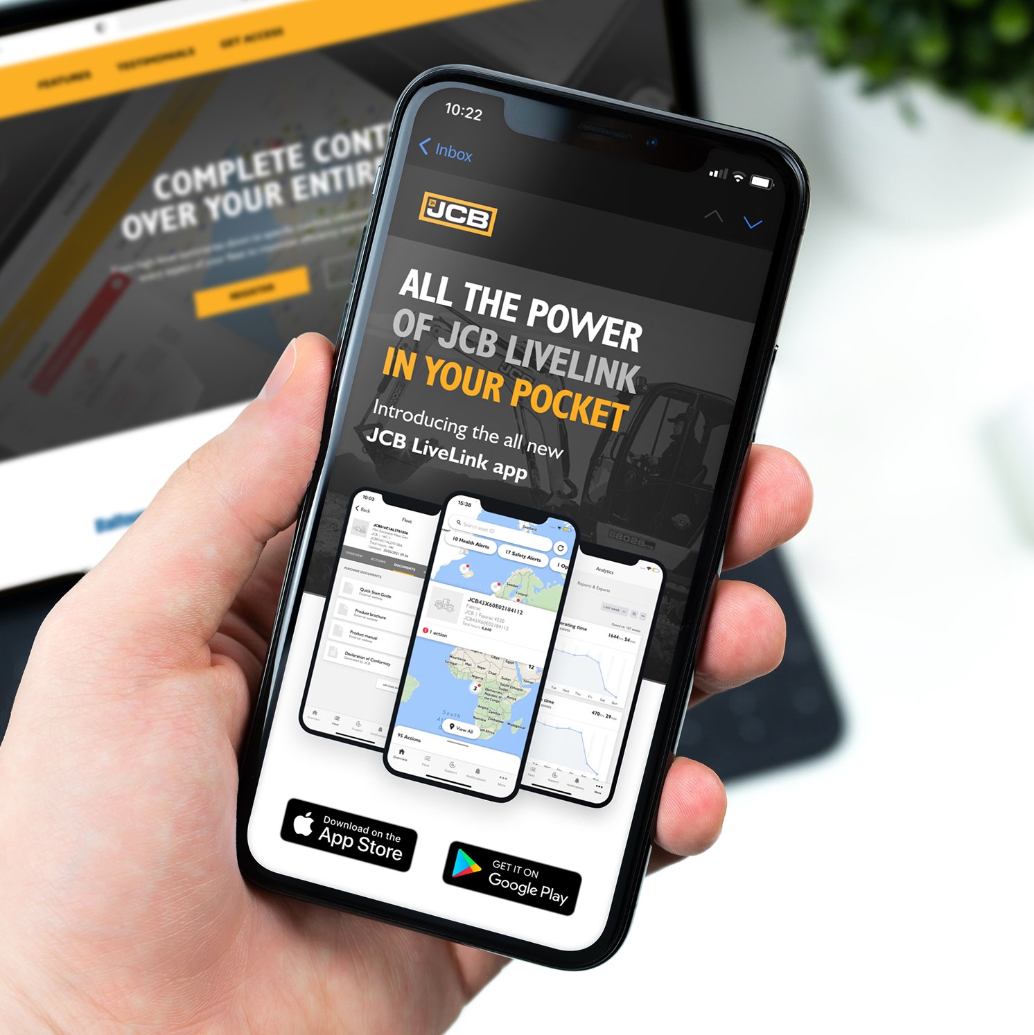 A hand holding a mobile phone displaying an advert for the JCB LiveLink app, featuring app interface screenshots and download options from the App Store and Google Play. A laptop with the JCB website is blurred in the background.